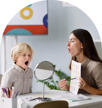 A speech therapist and a young girl sit on the floor, both pointing to their mouths while practicing speech exercises. The setting includes colorful therapy tools and toys, creating a supportive environment for children with apraxia of speech.
